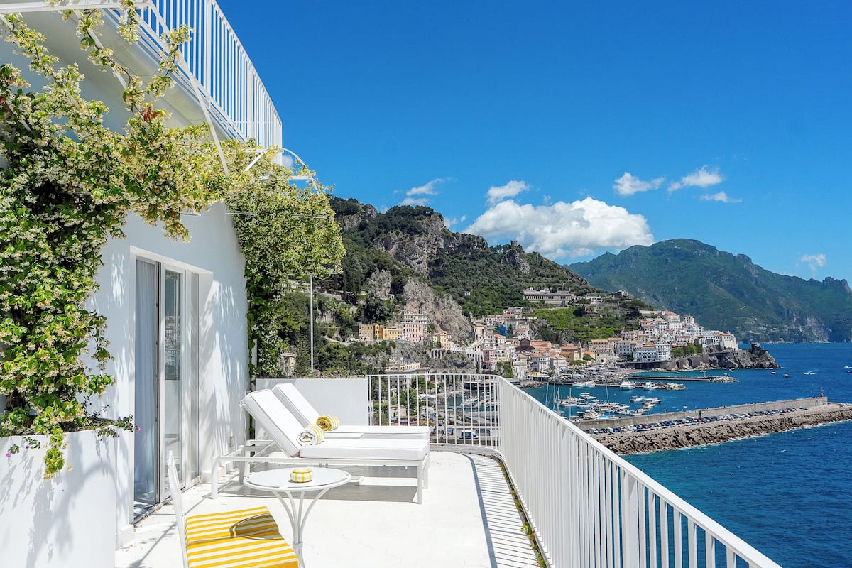 A balcony with white railings, a white chair, a yellow striped chair, and a small table with a yellow and white striped cushion.