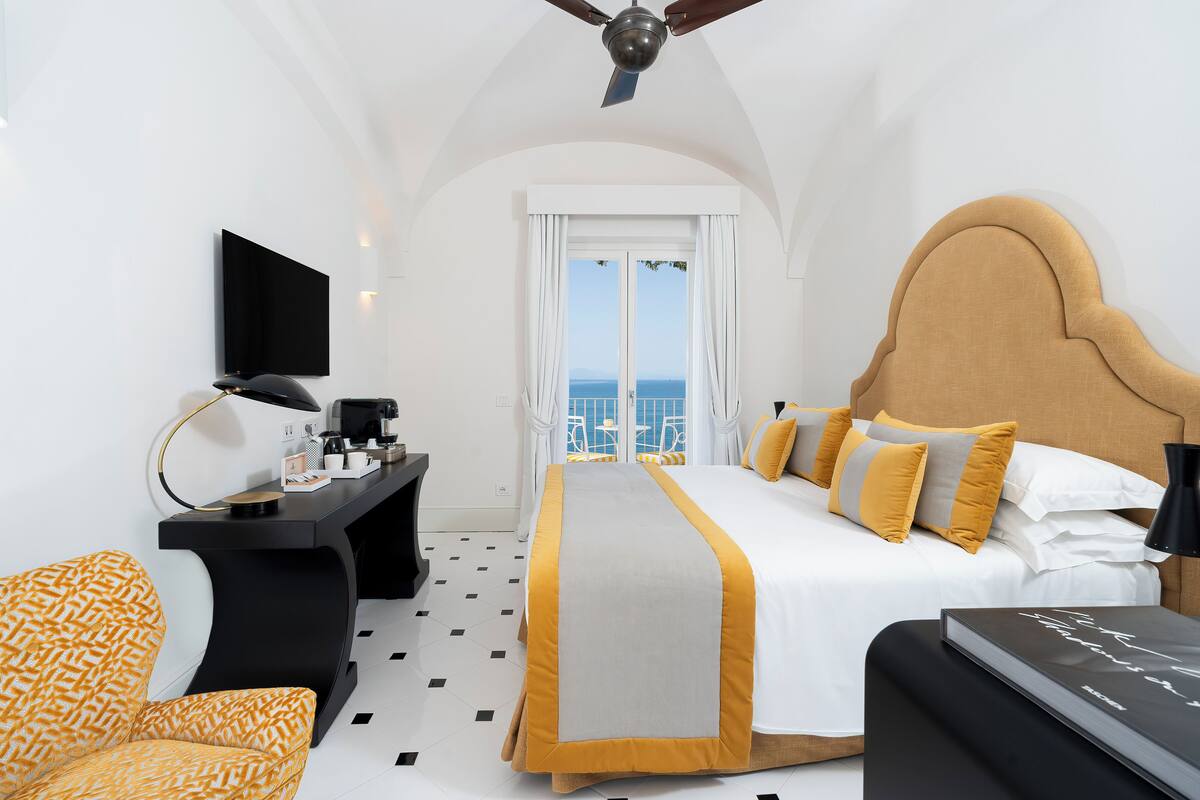 A hotel room with a bed, a desk, a chair, a television, and a view of the sea.