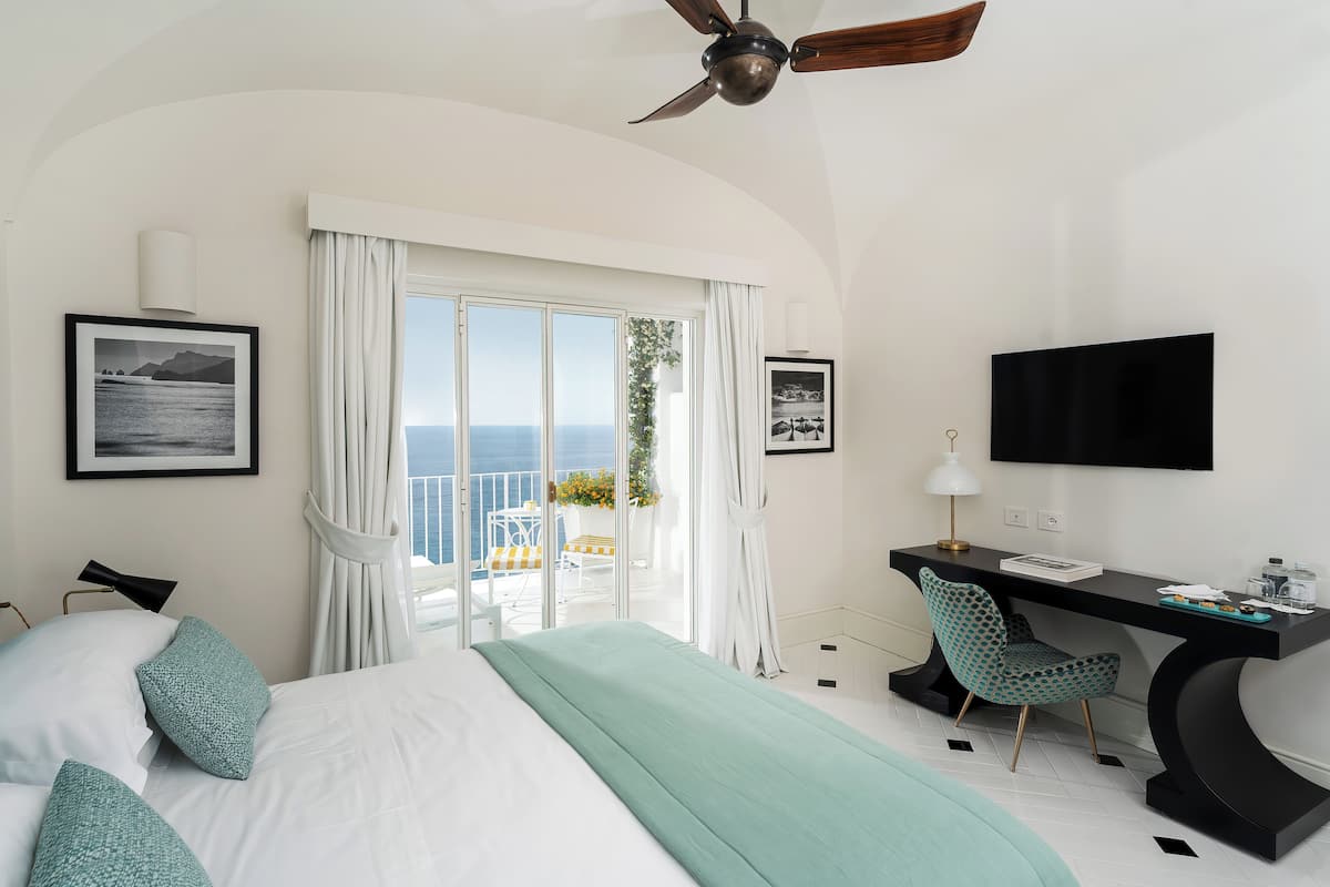 A hotel room with a bed, a desk, a chair, a TV, and a balcony with a view of the sea.
