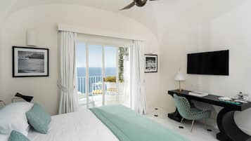 A hotel room with a bed, a desk, a chair, a TV, and a balcony with a view of the sea.