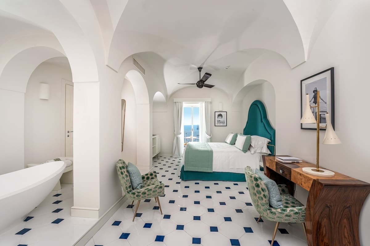 A bedroom with a large bed, a desk, a chair, a vanity, and a bathtub.