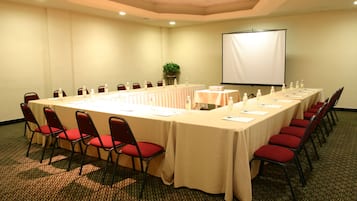 Un salón de conferencias con una larga mesa, sillas rojas, una pantalla de proyector y una planta en maceta.