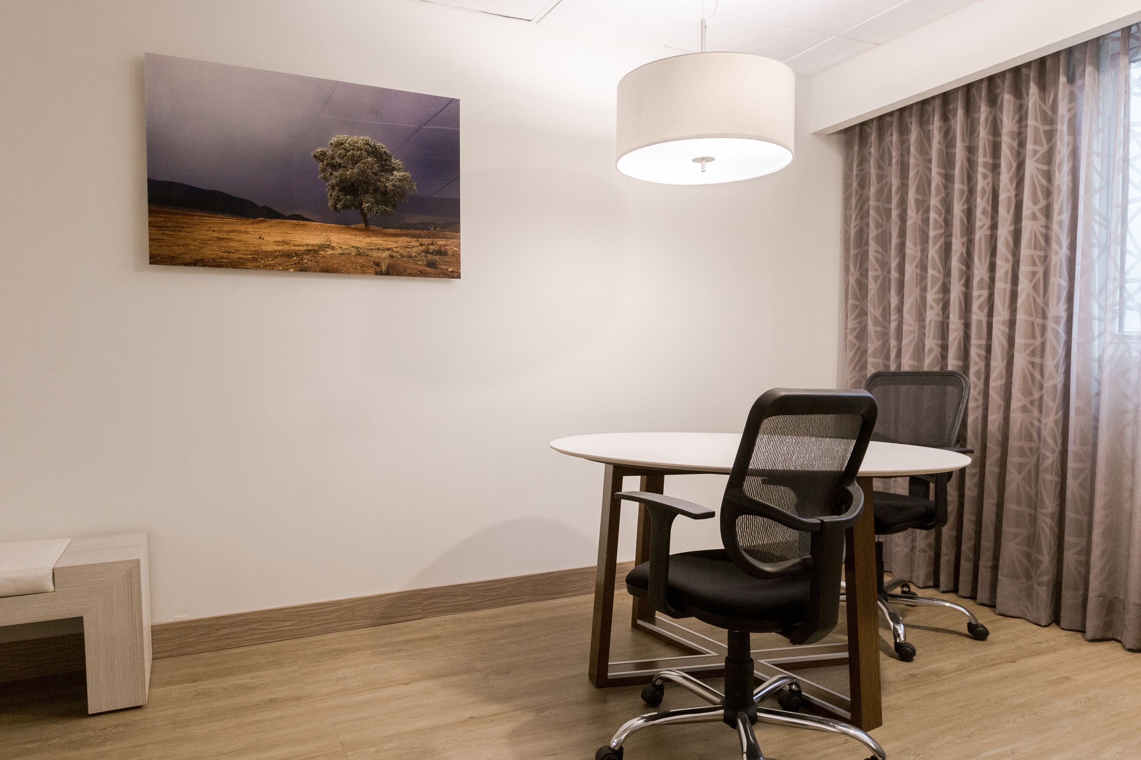 A conference room with a round table, two office chairs, a painting of a tree on the wall, a hanging light fixture, and a window with curtains.