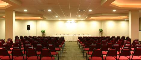 A conference room with rows of red chairs, a white table with microphones, and a speaker at the front.