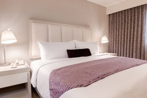 Habitación de hotel con una cama grande, cabecera blanca, dos mesitas de noche con lámparas, un teléfono y una ventana con cortinas.