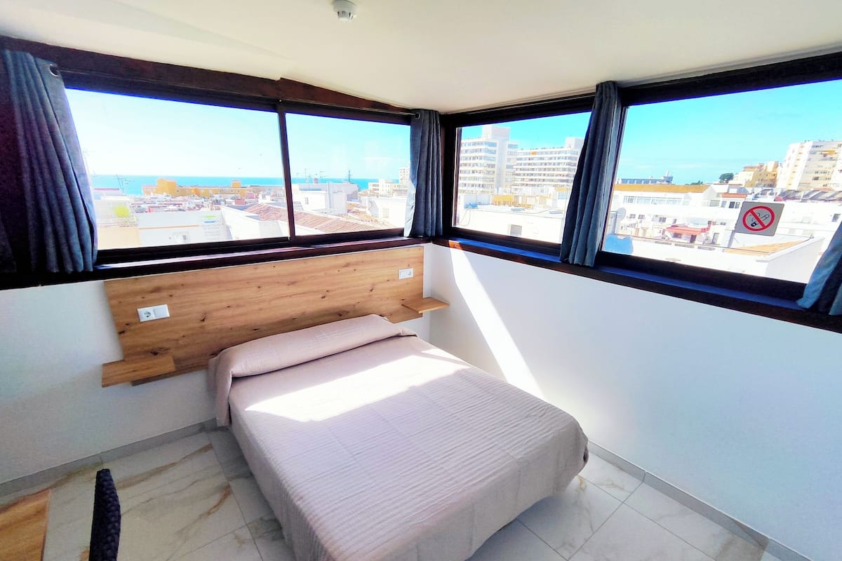 Een slaapkamer met een bed, een houten hoofdbord, grote ramen en uitzicht op de stad.