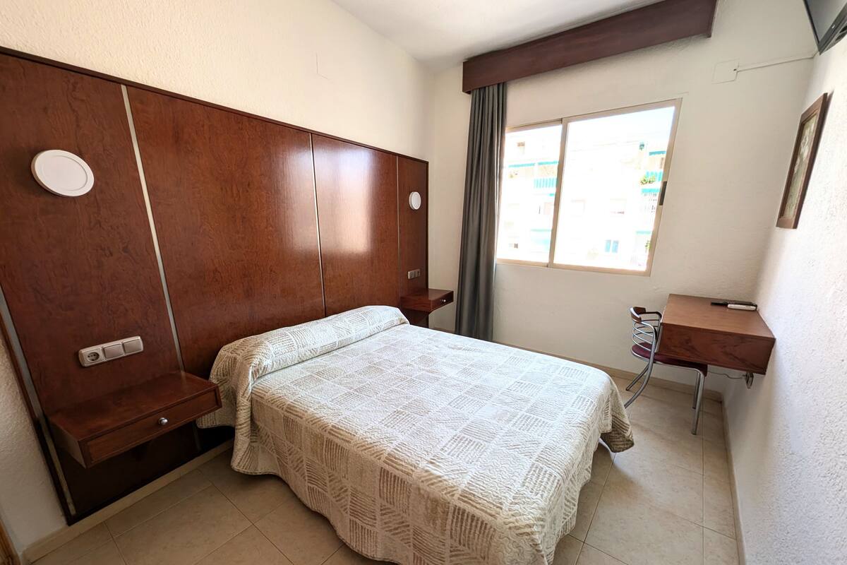 Een slaapkamer met een houten hoofdbord, een bed met een gebreide deken, een bureau met stoel en een raam met gordijnen.