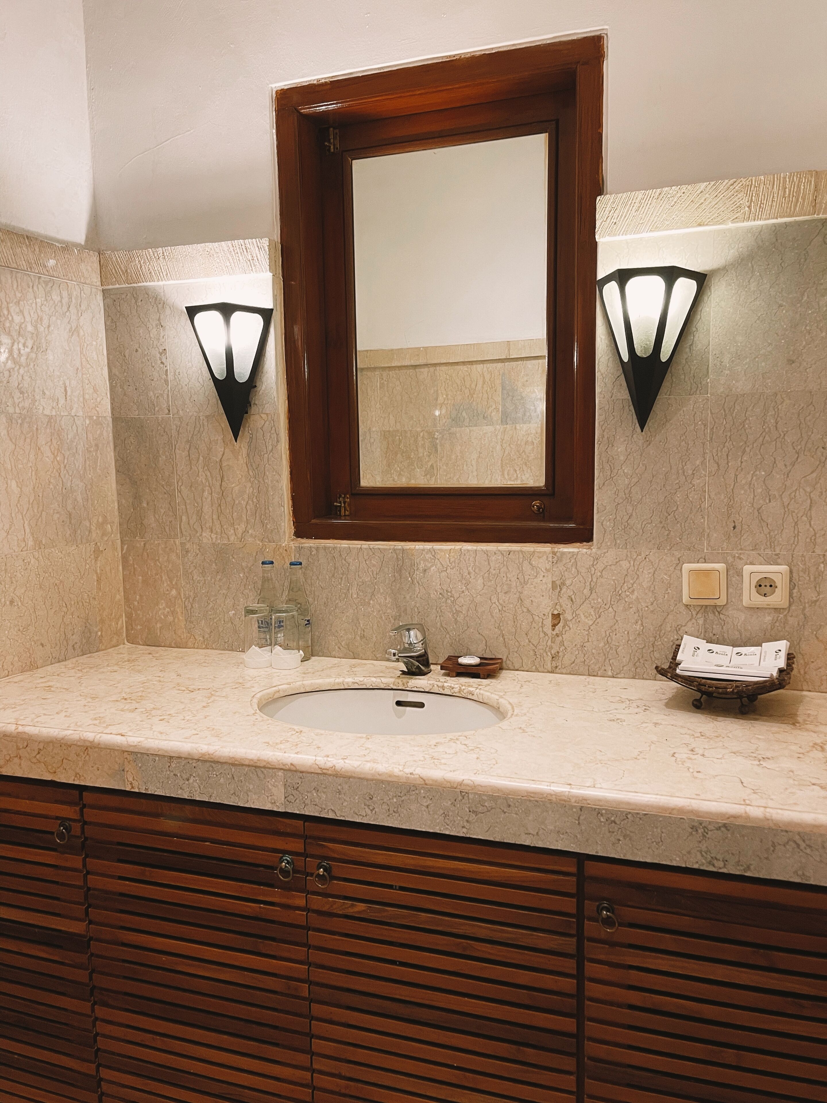 A bathroom with a marble sink, wooden cabinets, a mirror with a wooden frame, and two wall-mounted light fixtures.