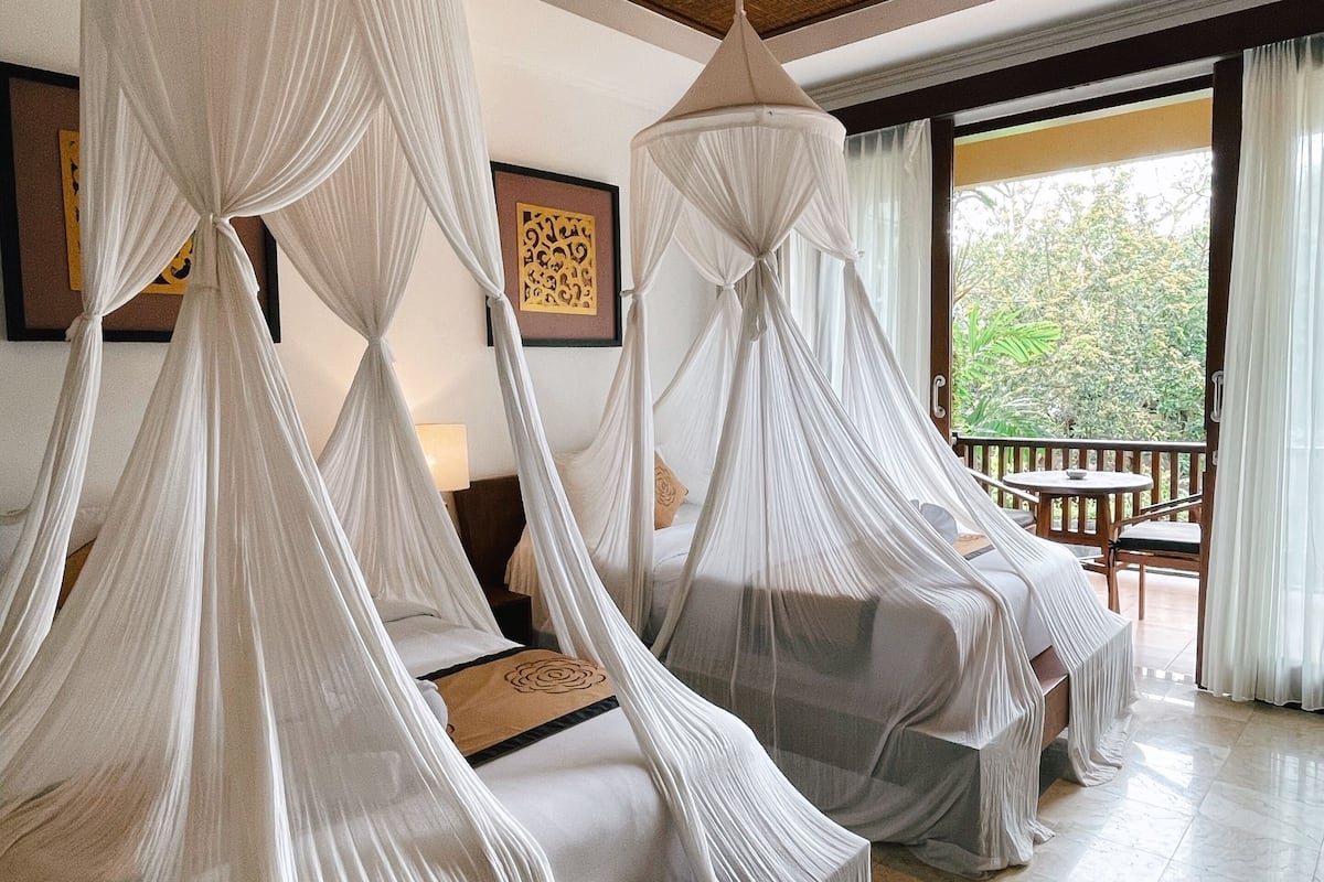 Habitación con dos camas cubiertas por doseles blancos, techo de madera y vistas a la vegetación exterior.