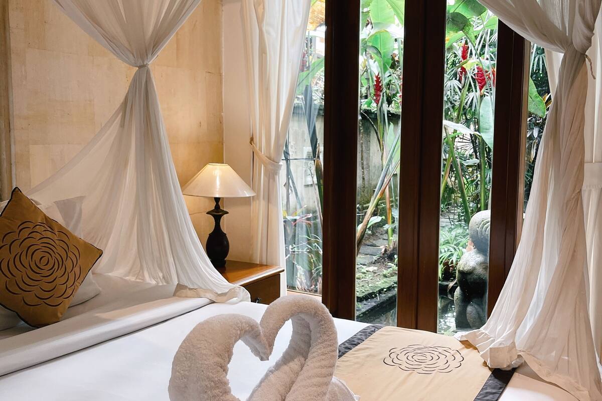 Habitación de hotel con cama, mesita de noche, lámpara y vista a un jardín a través de un ventanal.