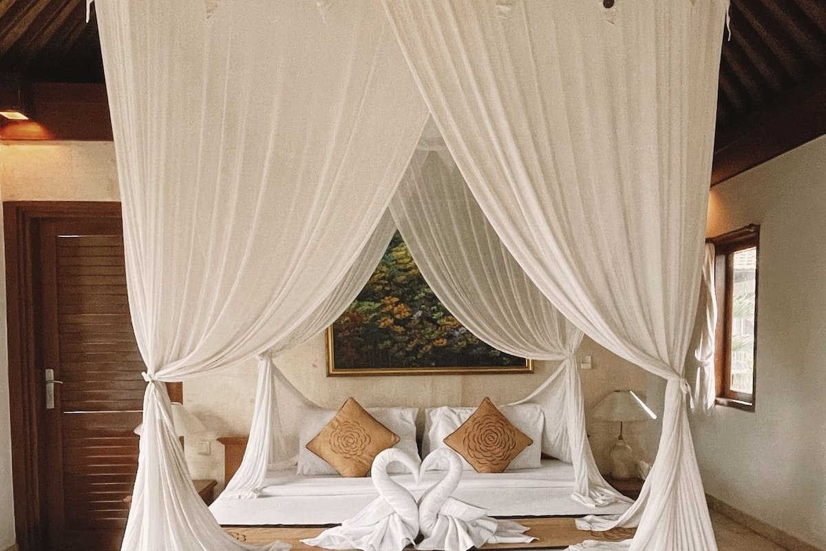 Una cama con dosel y cortinas, con una decoración de almohadas en forma de cisne, un cabecero de madera y un cuadro en la pared.