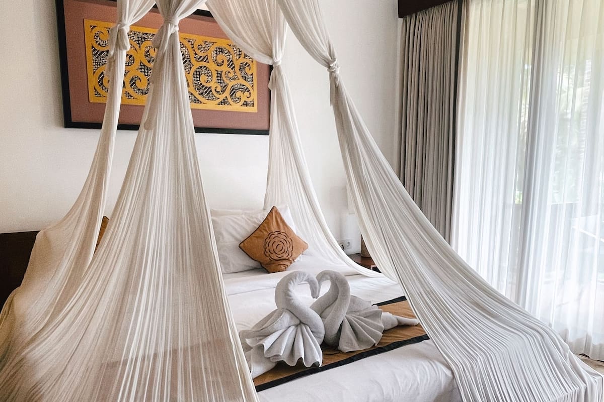 Un dormitorio con cama con dosel, cabecera de madera, un ventanal con cortinas translúcidas y una obra enmarcada en la pared.
