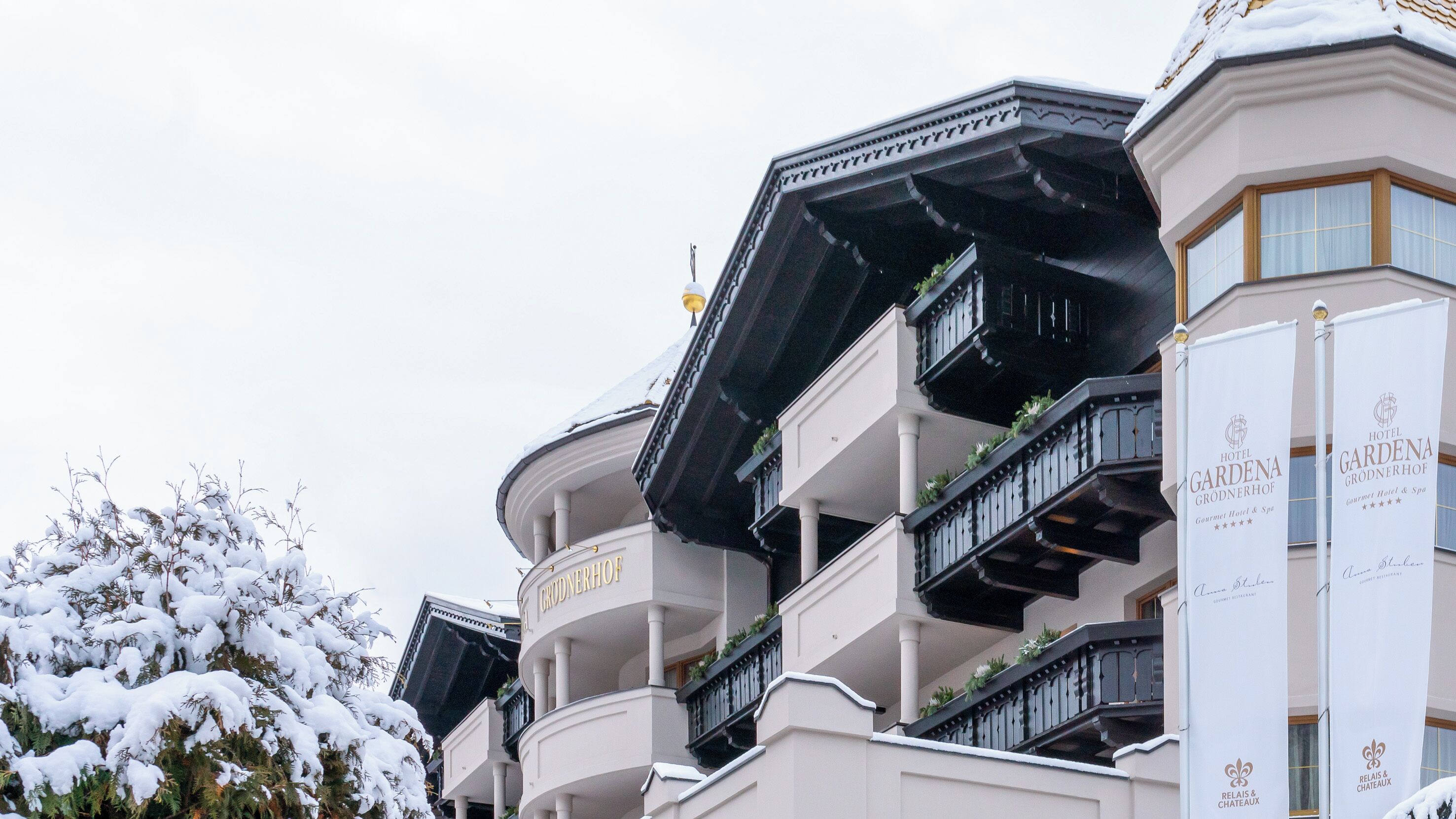 Un hotel de varios pisos con balcones, un árbol cubierto de nieve y señalización para Hotel Gardena y Relais & Chateaux.