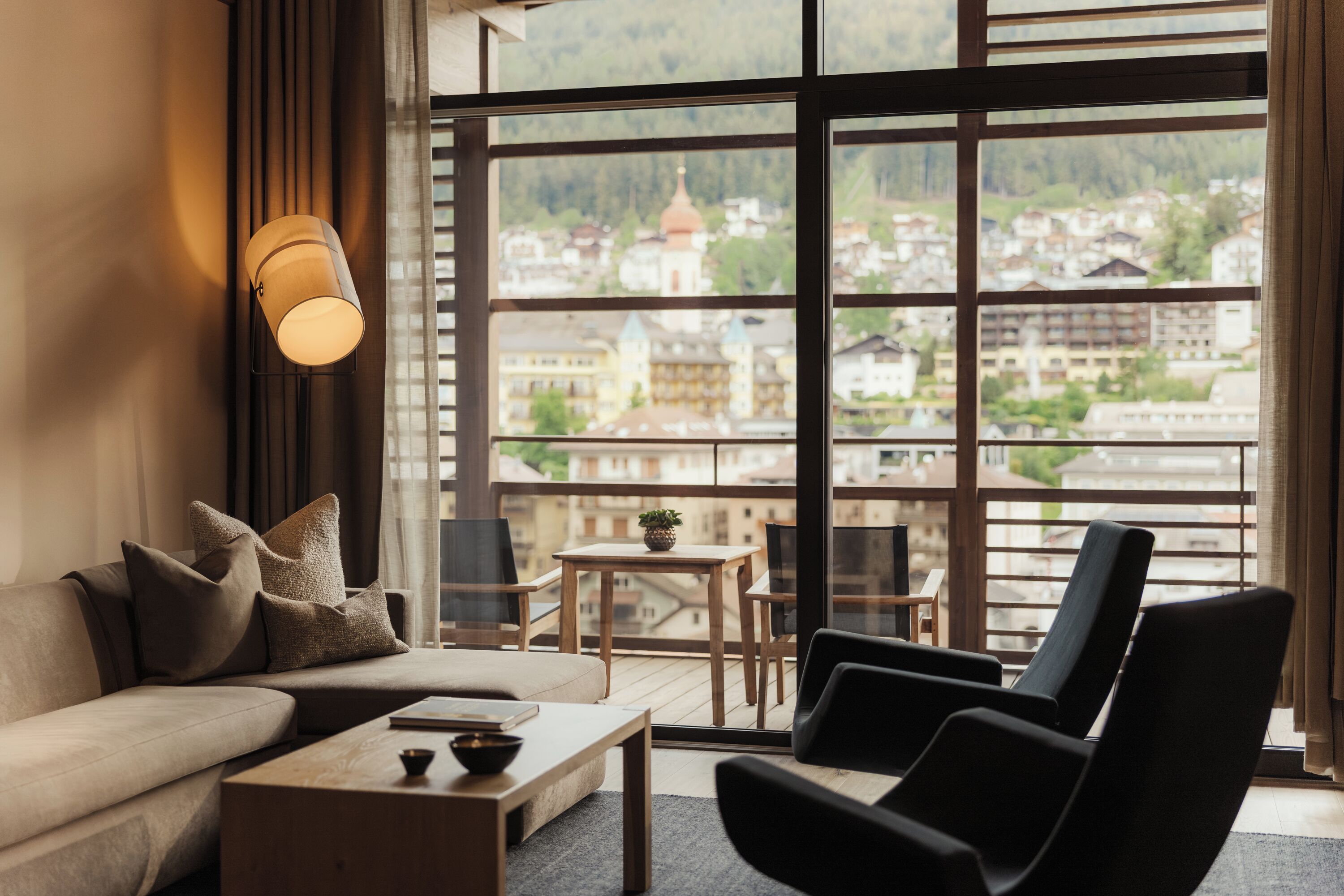 A modern living room with a sofa, armchair, coffee table, and a view of a cityscape through large windows.