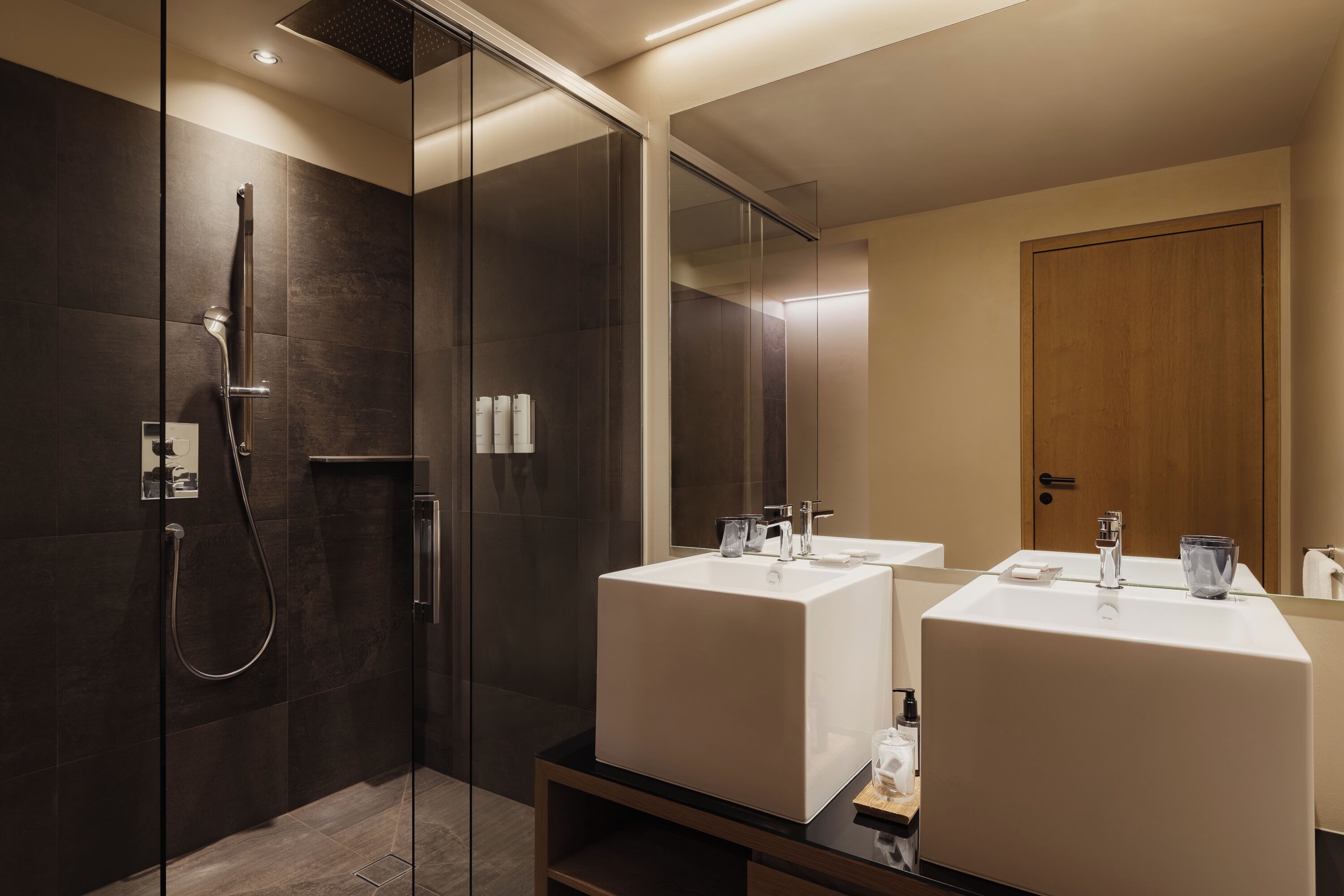 Ein modernes Badezimmer mit einer gläsernen Duschkabine, zwei Waschbecken und einer Holztür.