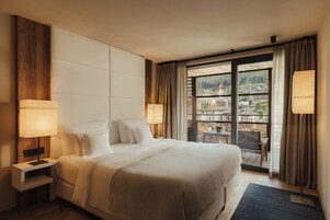 A hotel room with a large bed, two bedside lamps, a wooden headboard, a view of a town through a large window, and a balcony with a table and chairs.