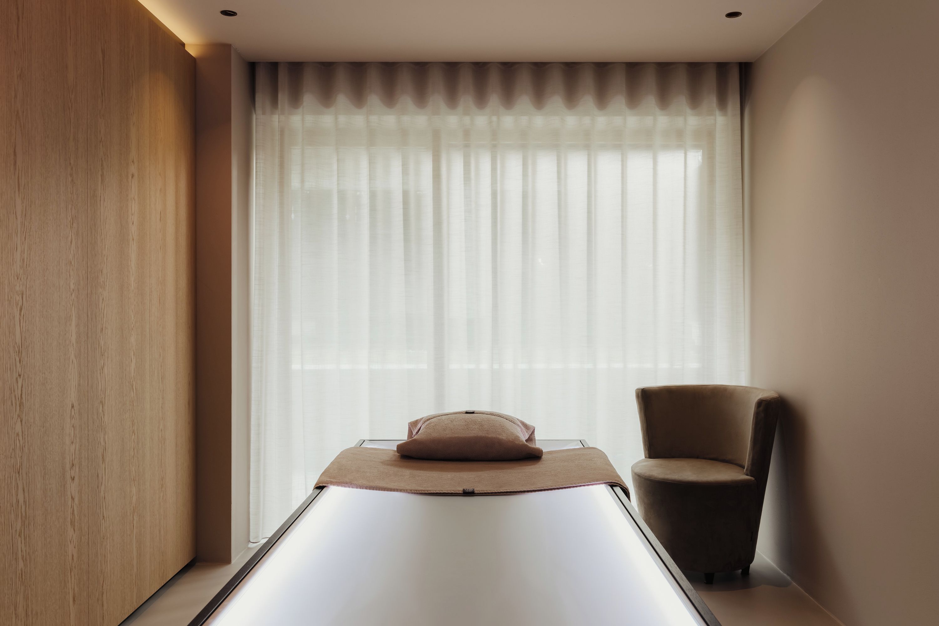 A room with a massage table, a chair, and a large window with sheer curtains.