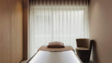 A room with a massage table, a chair, and a large window with sheer curtains.