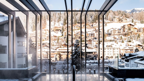 Une vue panoramique depuis une chambre d’hôtel située dans un immeuble de grande hauteur, avec de grandes fenêtres donnant sur un village enneigé et des montagnes.