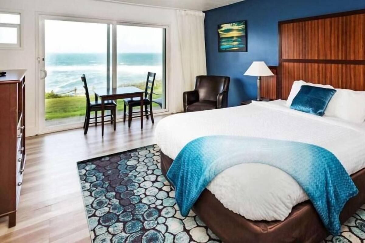 Une chambre avec un grand lit, une vue sur l’océan, une tête de lit en bois et un tapis à motifs de pierre.