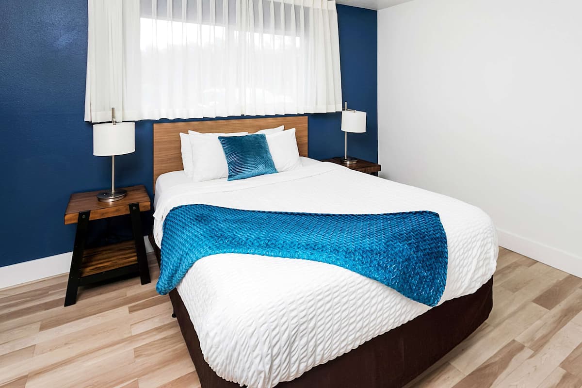 Un lit bien fait, recouvert d’un plaid bleu, deux tables de chevet avec des lampes et une fenêtre ornée de rideaux légers.