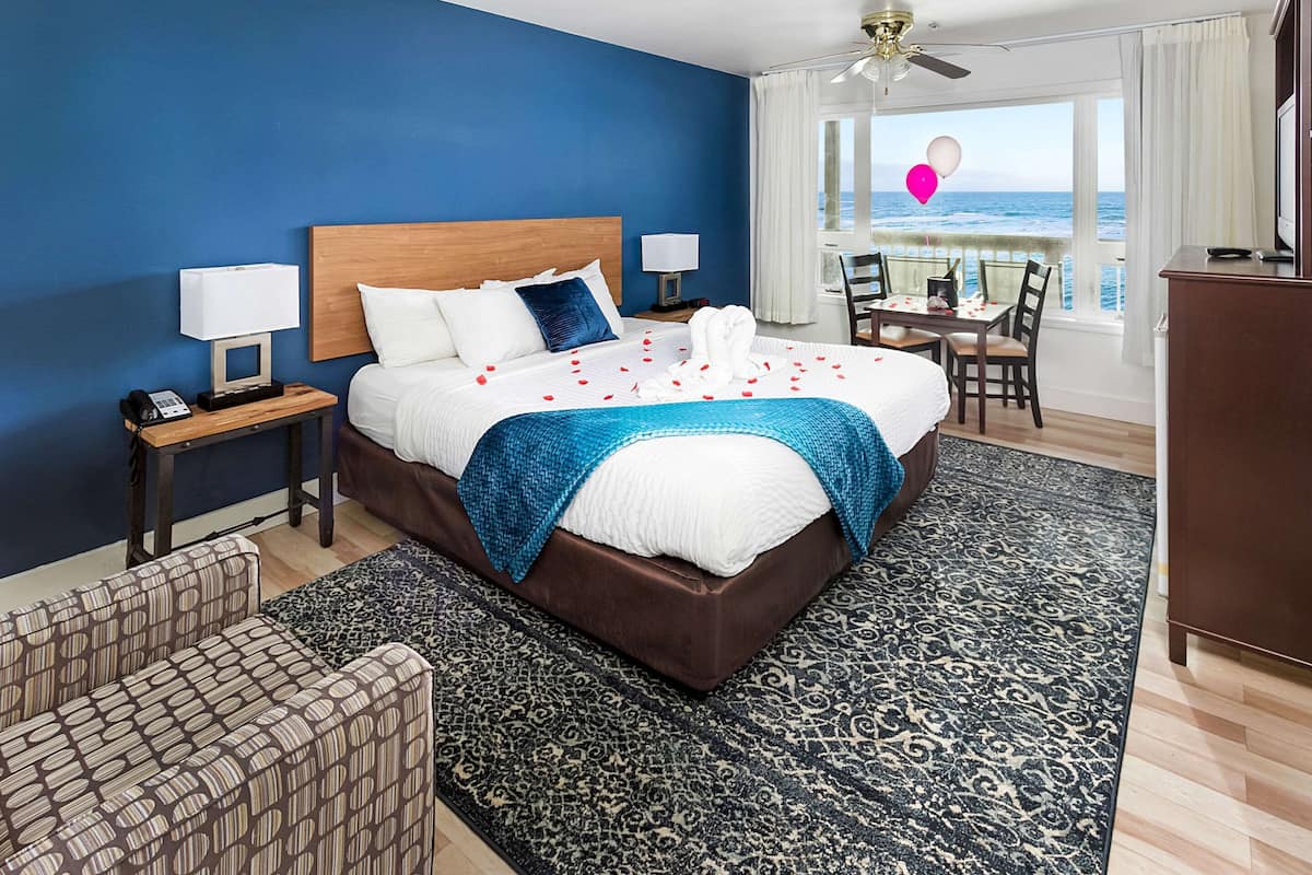Une chambre d’hôtel avec un grand lit, un bureau avec une chaise, une vue sur l’océan et un balcon avec une table et des chaises.