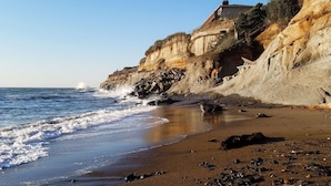 A coastal beach with a house on a cliff, waves crashing onto the shore, and a clear sky.