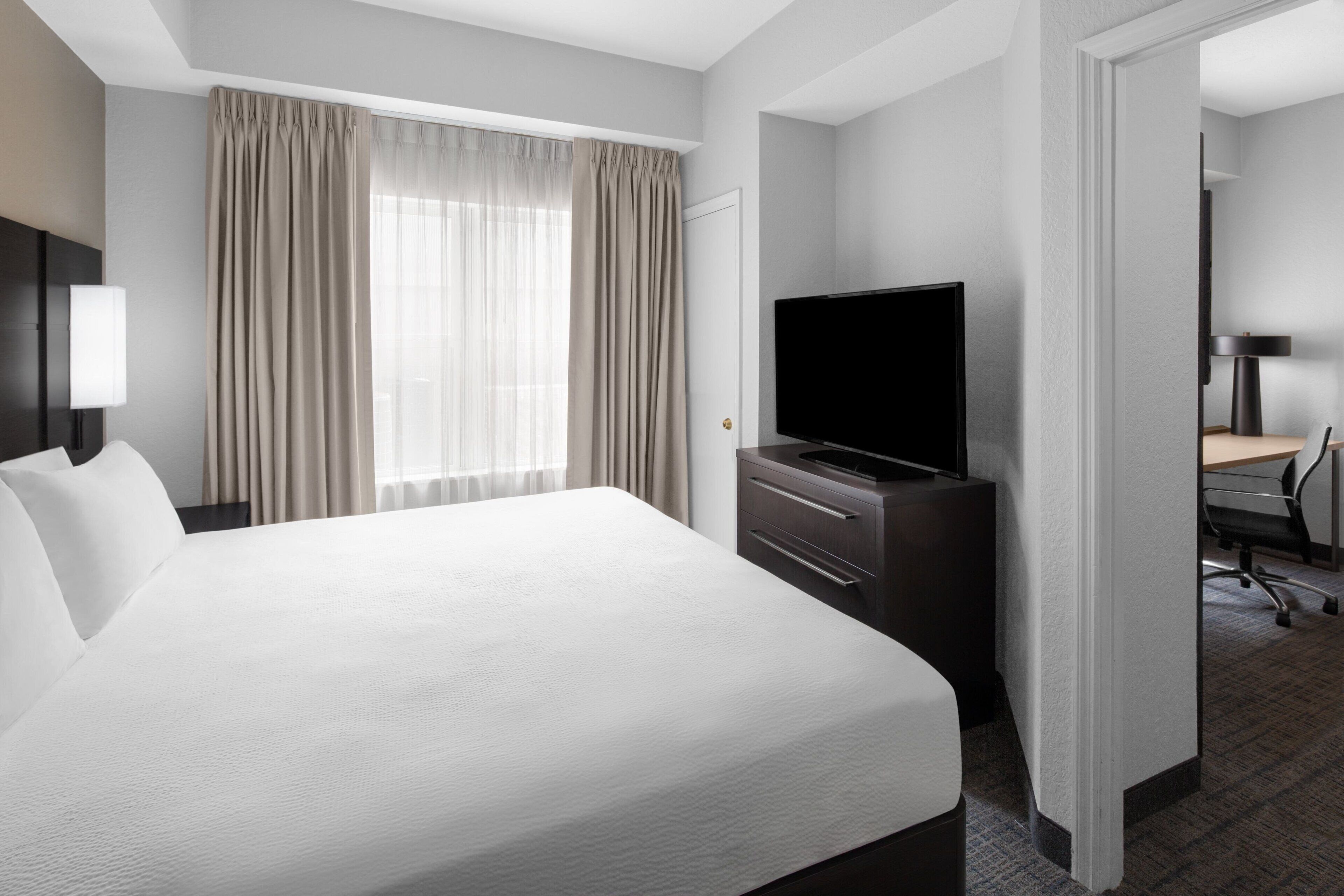 Una habitación de hotel con una cama grande, un televisor de pantalla plana, un escritorio con una silla y una ventana con cortinas.