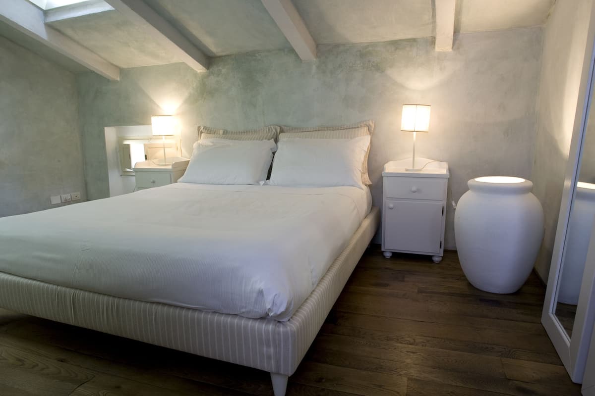 Ein Schlafzimmer mit einem großen Bett, zwei Nachttischen mit Lampen, einer großen Vase und einem Oberlicht.