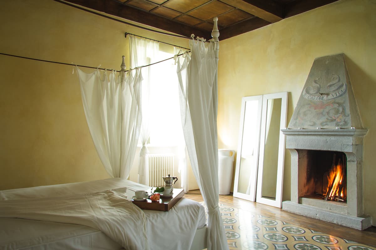 Ein Schlafzimmer mit einem Bett, einem Nachttisch mit Gegenständen darauf, einem Kamin und einem Fenster mit Vorhängen.