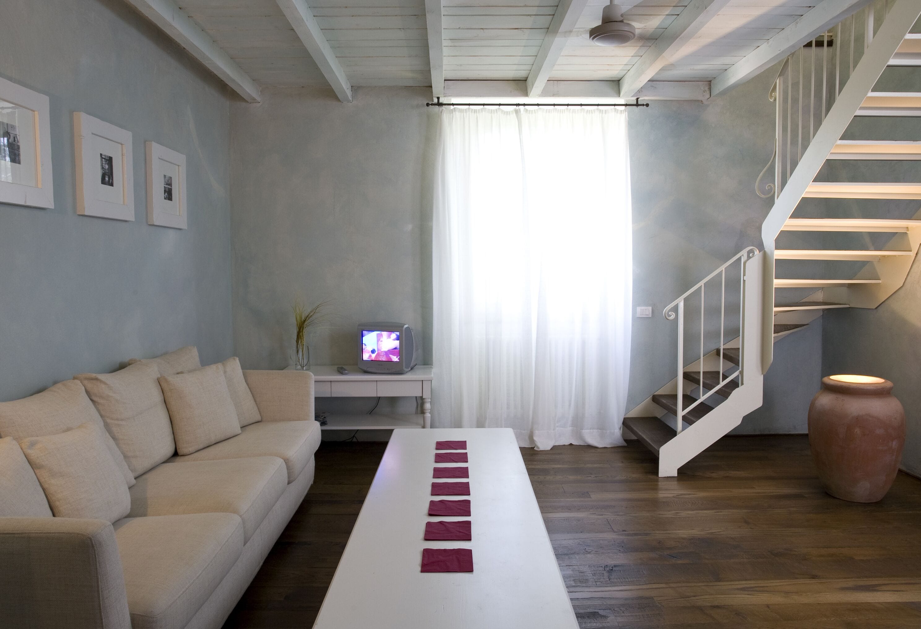 Una sala de estar moderna con un sofá, una mesa blanca alargada, una escalera y un televisor sobre una mesita.