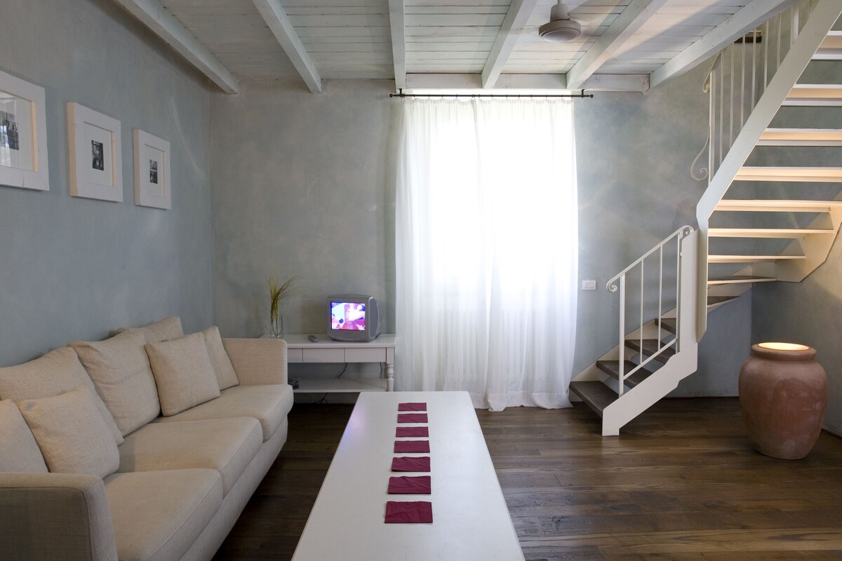 A modern living room with a sofa, a long white table, a staircase, and a television on a small table.