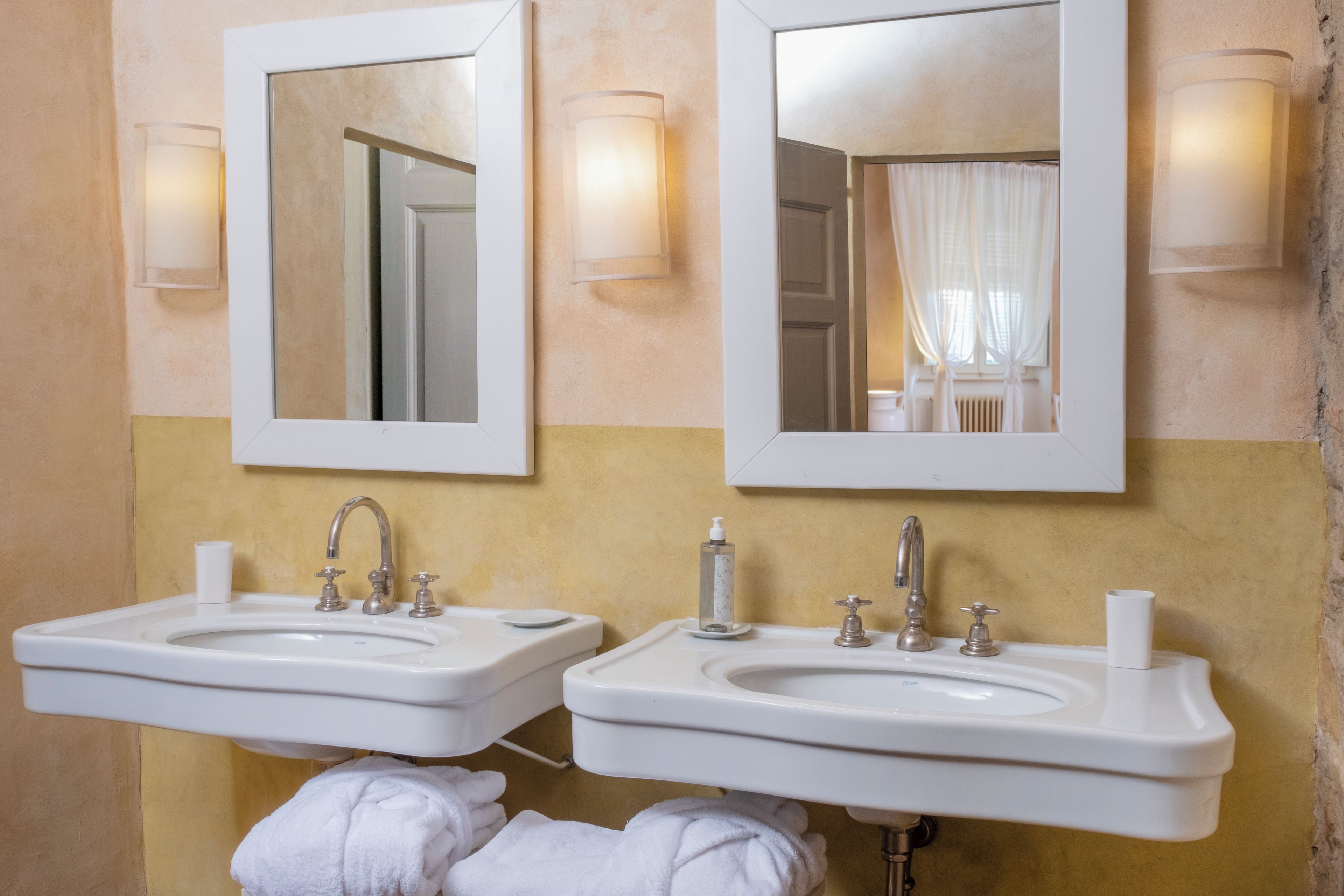 A bathroom with two sinks, each with a mirror above. There are towels on the floor and a bottle of hand soap on the sink.