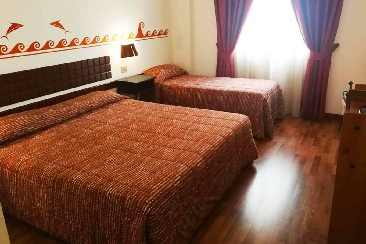 Une chambre d’hôtel avec deux lits, une table de chevet, une lampe et une fenêtre avec des rideaux violets.