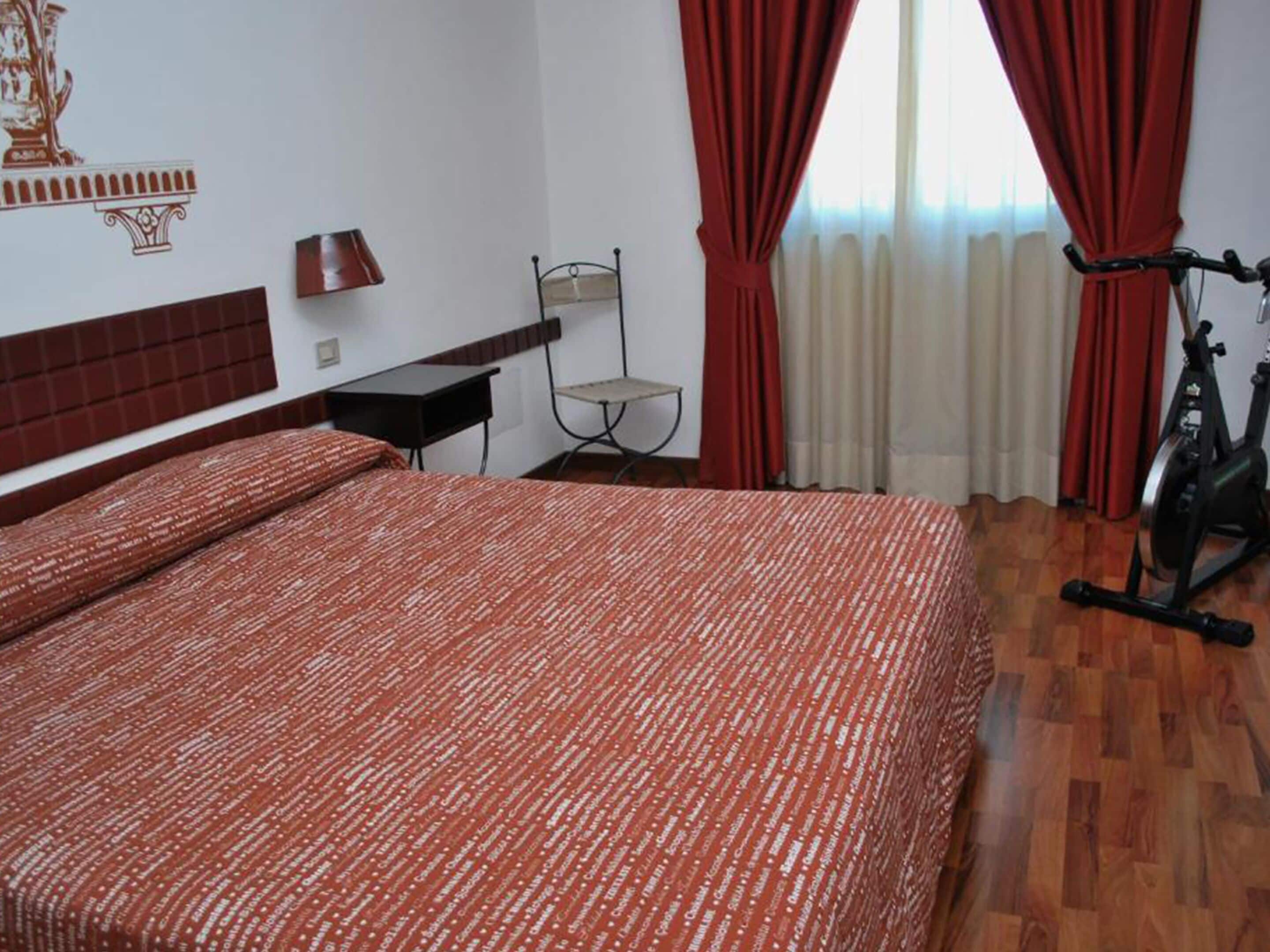 A hotel room with a large bed, a chair, a desk, a lamp, and an exercise bike.