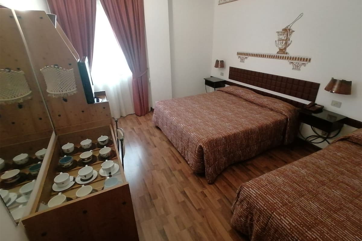 Une chambre d’hôtel avec un plancher en bois, un lit recouvert d’une couverture marron, une table de chevet avec une lampe et une étagère fixée au mur, sur laquelle sont posées des tasses et des soucoupes.