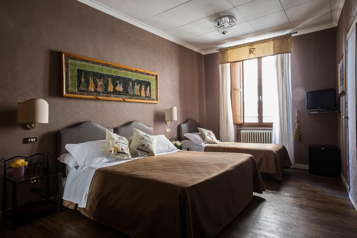 A hotel room with two beds, a large painting on the wall, a television, and a window with curtains.