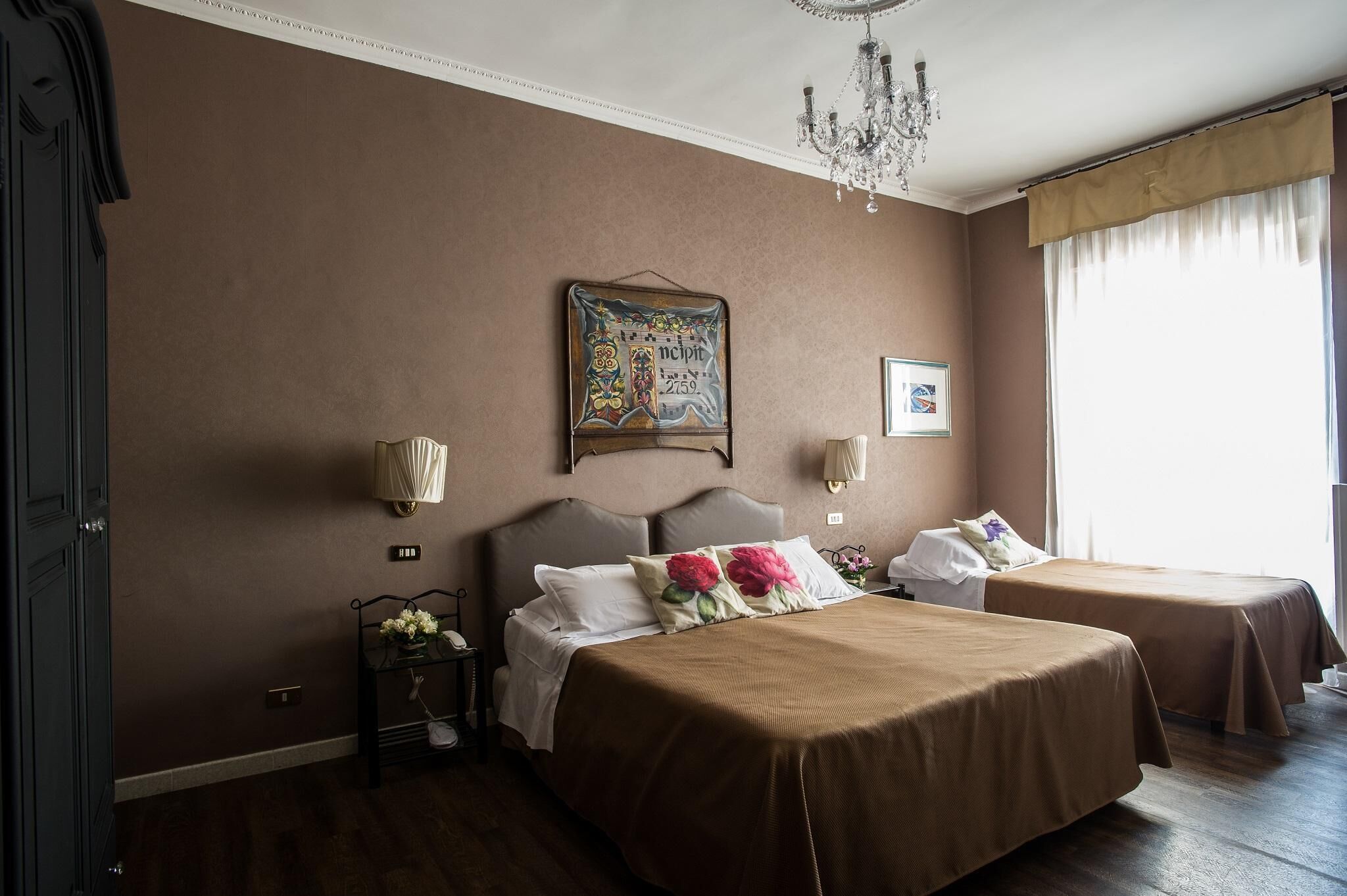 A hotel room with two beds, a chandelier, a framed picture on the wall, and a window with curtains.