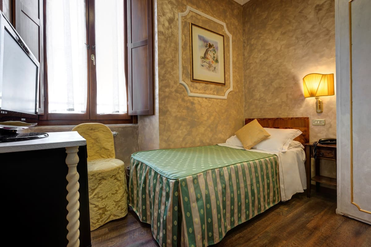 A room with a bed, a desk, a chair, a lamp, and a painting on the wall.