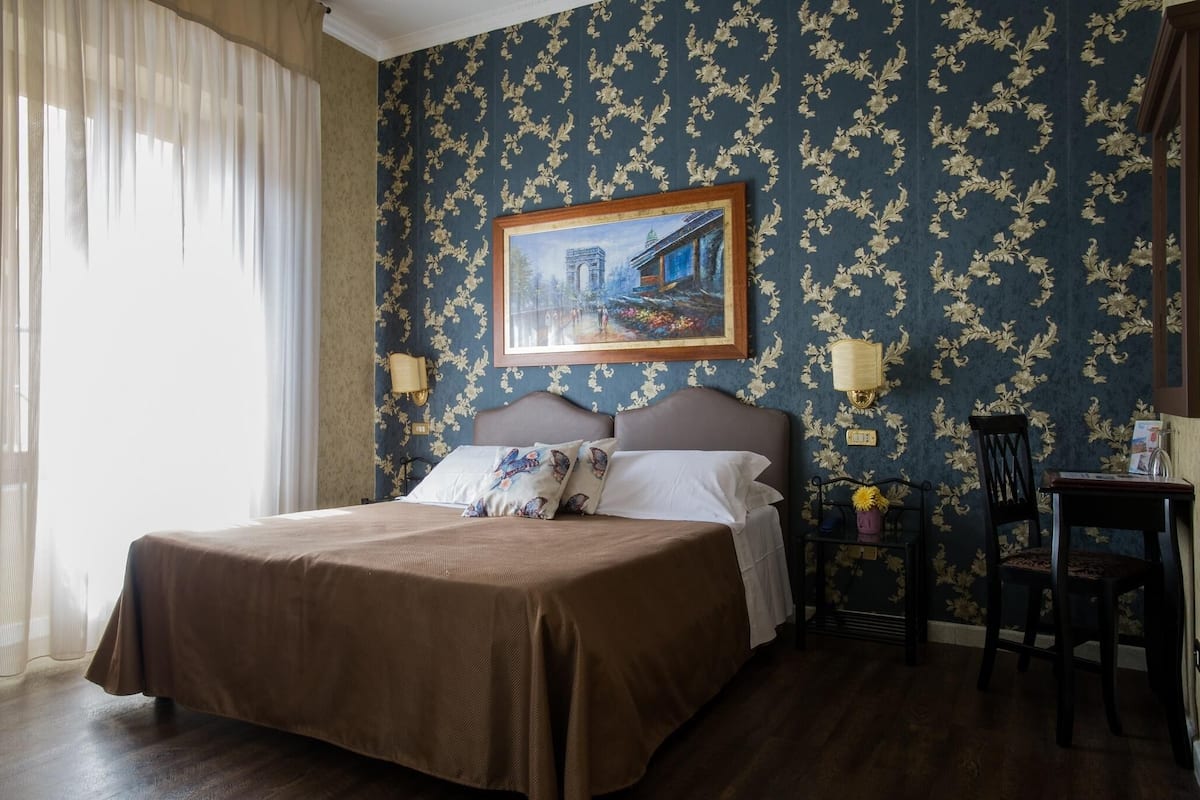 A hotel room with a large bed, a painting on the wall, and a desk with a lamp.