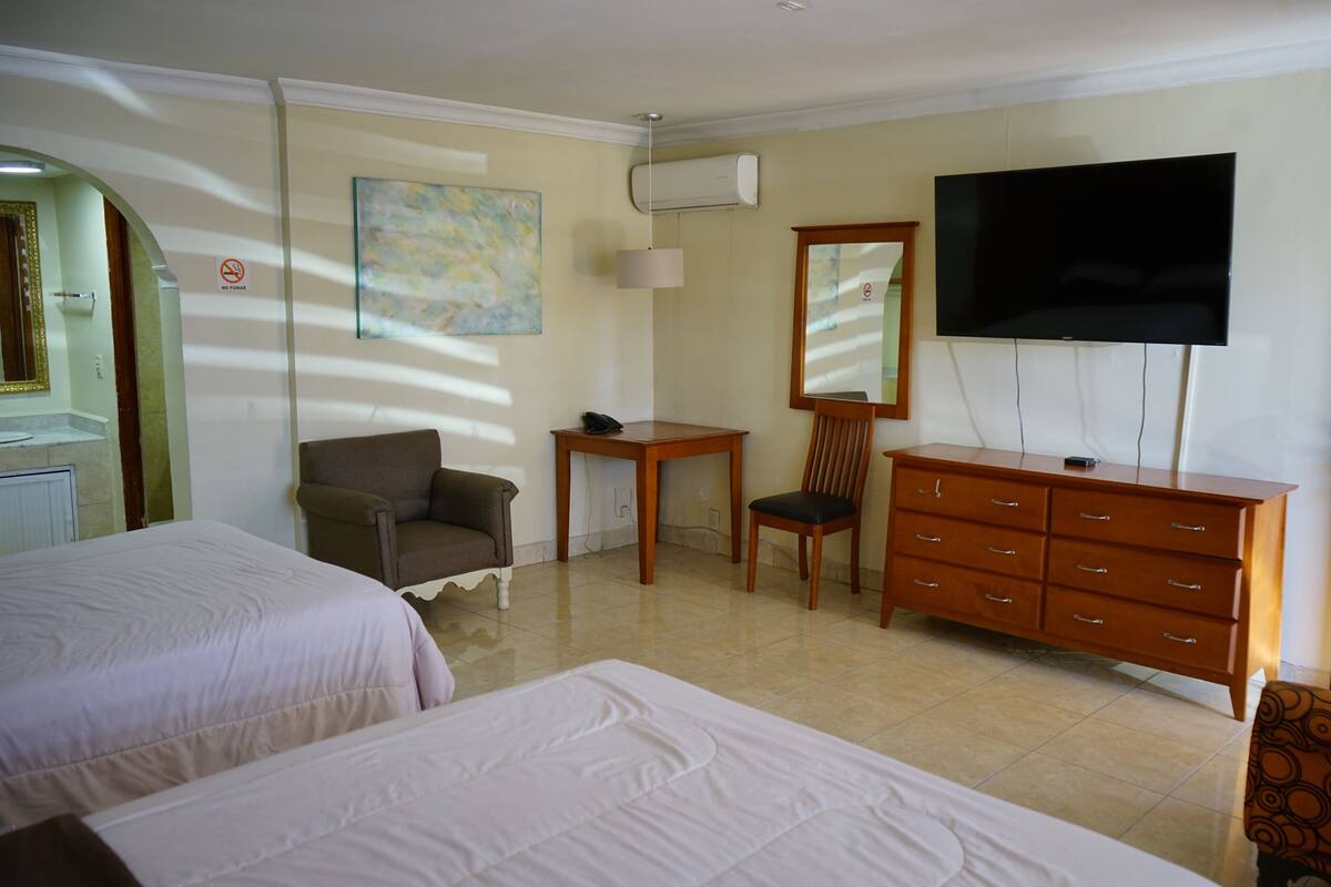 A hotel room with two beds, a desk, a chair, a dresser, a TV, and an air conditioning unit.