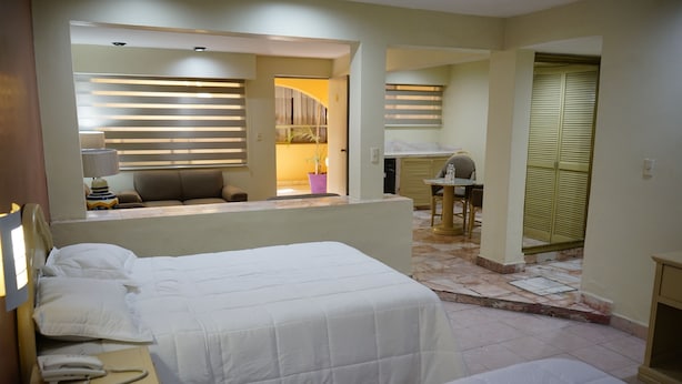 A hotel room with a bed, bedside table, lamp, and a view of a kitchen area. - Hotel Posada del Sol Inn (Torreón)