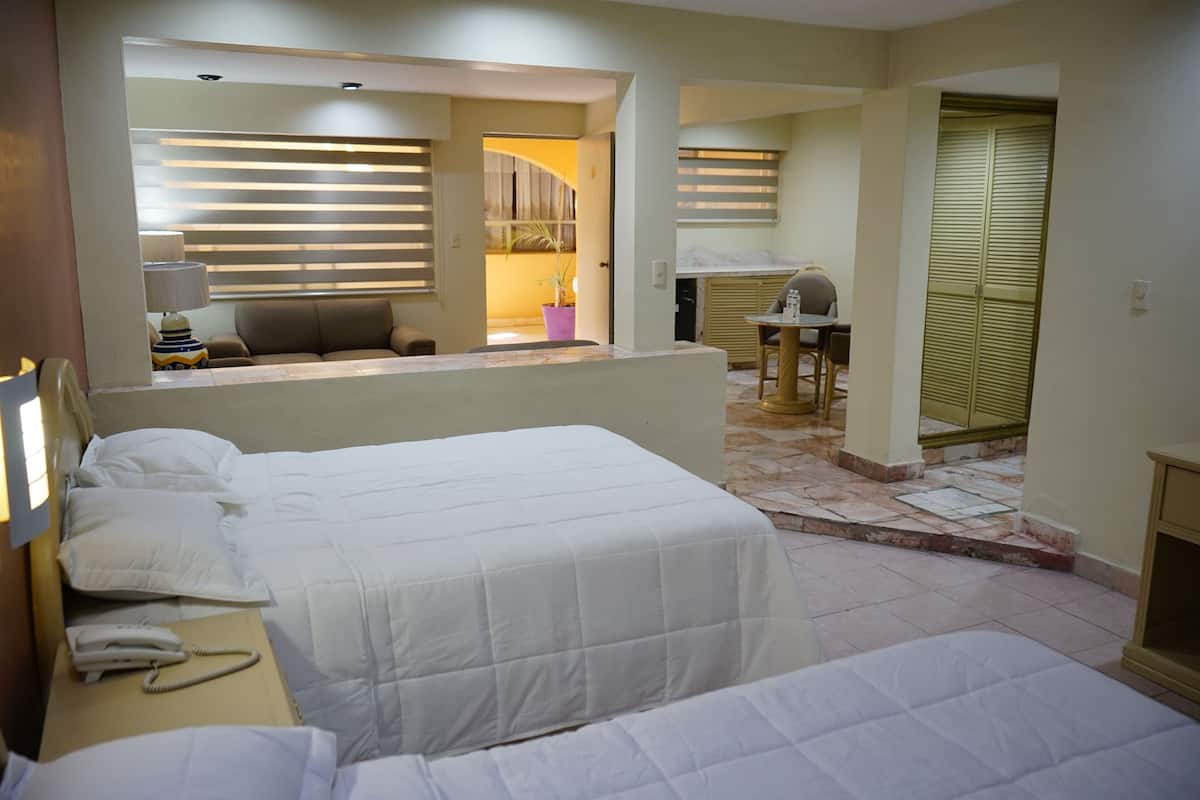 A hotel room with a bed, bedside table, lamp, and a view of a kitchen area.