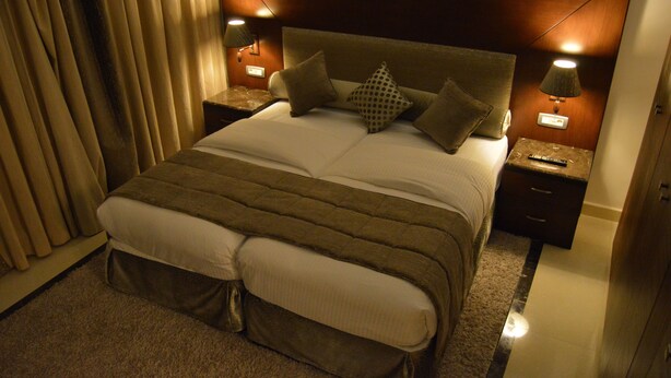 A neatly made bed with a brown headboard, two bedside tables with lamps, and a large window with curtains. - Markazia Suites (Beirut)