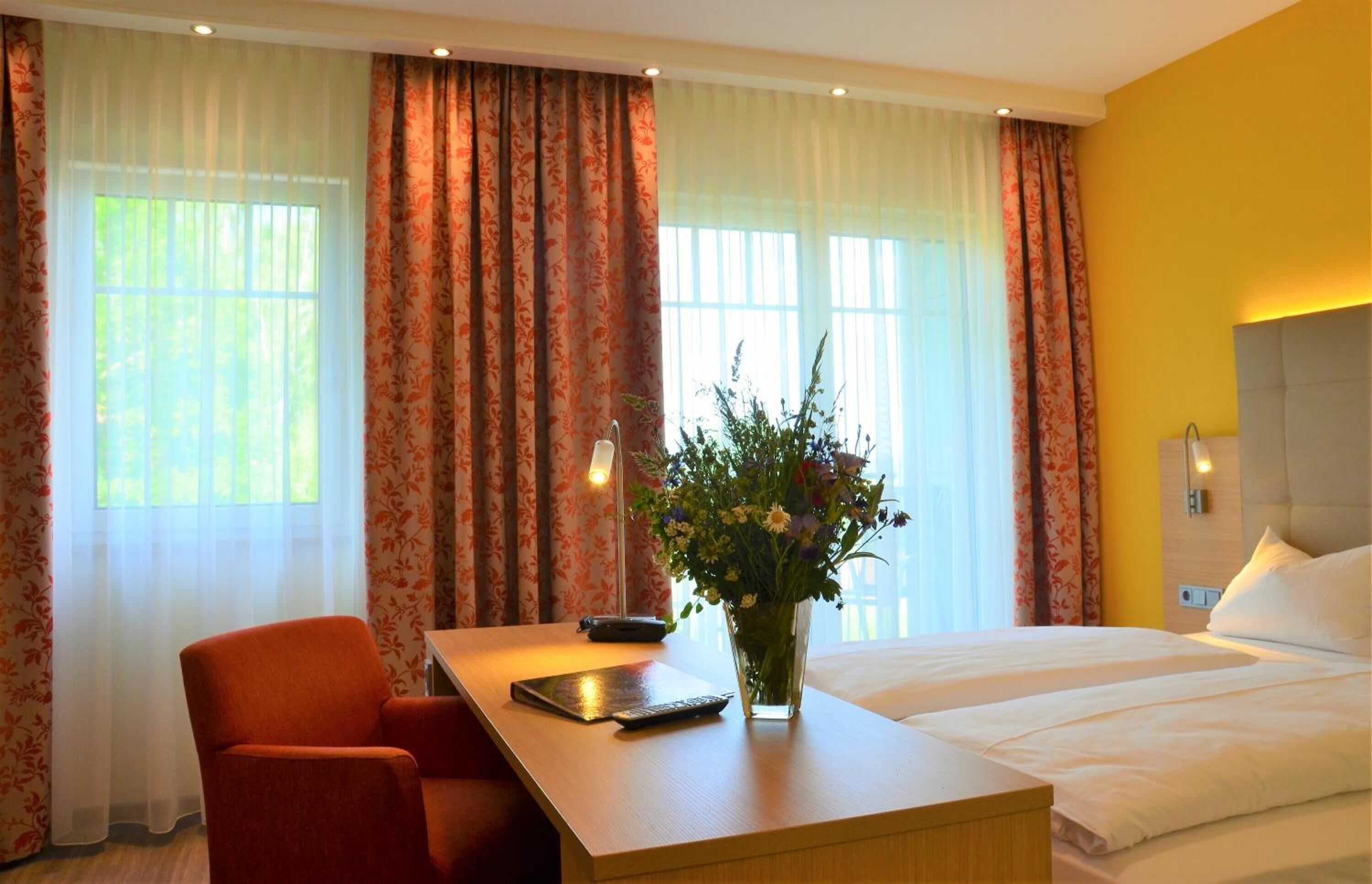 A hotel room with a bed, a desk with a chair, a vase of flowers, and curtains.
