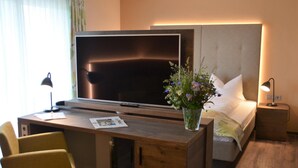 Habitación de hotel con escritorio de madera, televisor de pantalla plana, cama con ropa blanca, mesita de noche con lámpara y un florero con flores.