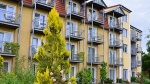 Un edificio residencial de varios pisos con balcones, un árbol siempreverde destacado y varios arbustos en primer plano.