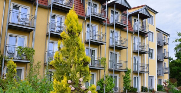 A multi-story residential building with balconies, a prominent evergreen tree, and a variety of shrubs in the foreground. - Hotel Bergwirt (Herrieden)