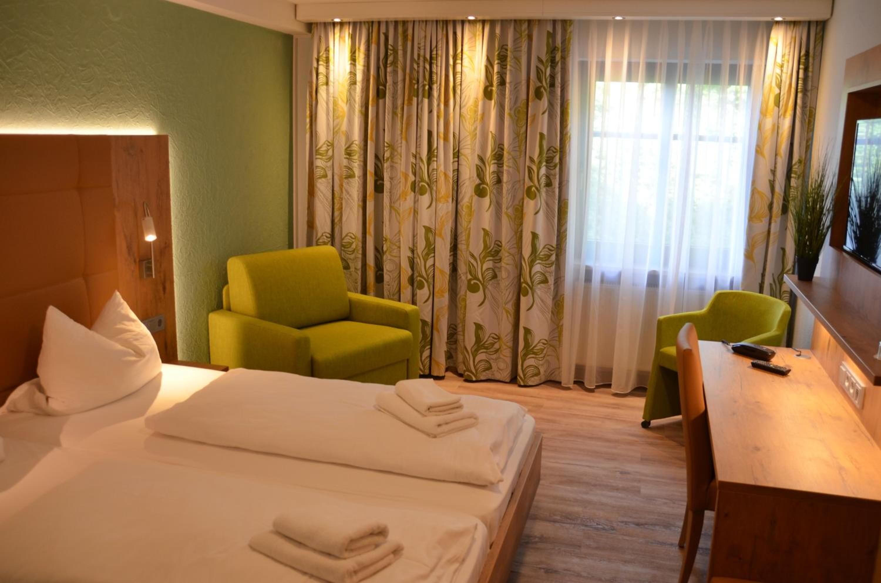 A hotel room with a bed, two green armchairs, a wooden desk, and a television.
