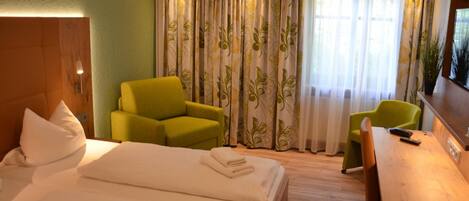 A hotel room with a bed, two green armchairs, a wooden desk, and a television.